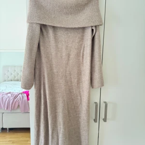 Beige stickad offshoulder långklänning - Mysig beige stickad långklänning med bred offshoulder-krage och långa ärmar. Klänningen har en rak och avslappnad passform, perfekt för dig som gillar en enkel men ändå trendig look. Materialet är mjukt och värmande, perfekt för kyligare dagar.