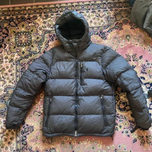 Grå dunjacka från Polo Ralph Lauren - Säljer en grå dunjacka från Polo Ralph Lauren med huva och dragkedja framtill. Jackan har tre fickor med dragkedja och den klassiska Polo-loggan broderad på bröstet. Perfekt för kalla dagar och riktigt skön att bära. Defekter en liten lagning ett litet hål samt att en dragkedja saknas