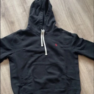 Svart hoodie från Polo Ralph Lauren - Svart hoodie från Polo Ralph Lauren med klassisk röd broderad logga på bröstet. Tröjan har huva med vita snören och en stor magficka. Tillverkad i mjuk bomull, perfekt för chill dagar. Snygg och enkel att matcha med allt.