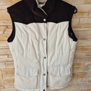 Vit och brun dunväst från Tindra - Snygg dunväst från Tindra i vit och mörkbrun färg med manchesterdetaljer upptill. Västen har tryckknappar framtill, två stora fickor och ett rutigt innerfoder med dragkedja. Perfekt för lager-på-lager och chill stil. Aldrig använd.