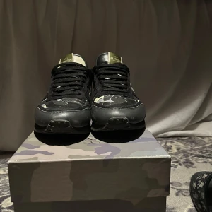 Valentino Garavani Rockrunner sneakers - Snygga Valentino Garavani Rockrunner sneakers i svart och grått med camouflage-mönster. Skorna har svarta snören, rund tå och detaljer i skinn och textil. Orange detalj på hälen ger extra edge. Kommer med originalkartong i matchande camo. 