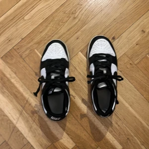 Nike Dunk Low svart/vit sneakers - Säljer ett par Nike Dunk Low sneakers i klassisk svart och vit färg. Skorna har svart sula, vit tåbox med perforeringar och svarta snören. Ikonisk Swoosh-logga på sidan och låg silhuett som passar till streetwear. Tillverkade i skinn med platt sula. Har också slitningar på snörena