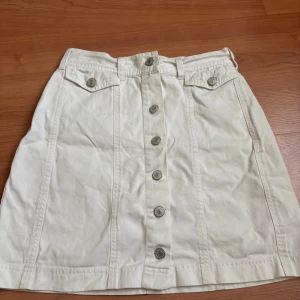 Vit kortkjol med knappar Sinclair KappAhl - Snygg vit kortkjol från Sinclair by KappAhl med raka linjer och coola silverfärgade knappar framtill. Kjolen har två fickor med lock och bälteshällor i midjan. Tillverkad i bomull, perfekt för en fräsch och stilren look.