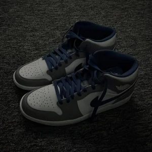 Nike Air Jordan 1 Mid Blå/Vit - Snygga Nike Air Jordan 1 Mid sneakers i blått och vitt. Skorna har klassisk snörning, perforerad tå och ikoniska Jordan Wings-loggan på sidan. Perfekt för dig som vill ha en stilren och sportig look med streetkänsla.