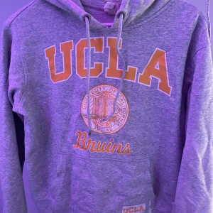Grå UCLA hoodie med tryck - Säljer en grå hoodie från UCLA med stort orange och vitt tryck framtill och snörning i huvan. Klassisk känguruficka och mjukt material, perfekt för chill dagar eller plugget. Bruins-logga på magen och liten patch nertill.
