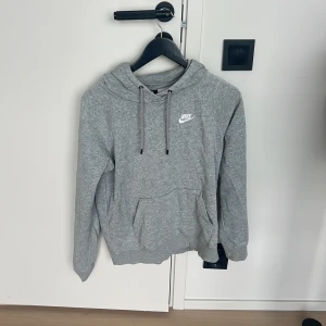 Grå hoodie från Nike med huva - Säljer en grå hoodie från Nike med klassisk logga på bröstet. Tröjan har huva med snörning och en stor magficka. Perfekt för chill dagar eller när du vill ha en enkel och stilren look.