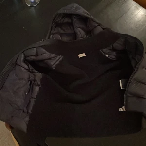 Svart Moncler pufferjacka med huva - Svart pufferjacka från Moncler med stickade ärmar och vadderad kropp. Jackan har huva, dragkedja framtill och silverfärgade knappar med Moncler-logga. Klassisk logotyp på bröstet och snygg, sportig look. Perfekt för kyliga dagar. Kvitto finns,pris kan diskuteras 