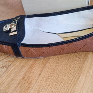 Michael Kors Hamilton moccasins - Hej Snygga Michael Kors Hamilton moccasins i brunt och svart skinn med quiltad struktur och guldfärgade MK-detaljer framtill. Skorna har rund tå och platt sula, perfekt för en stilren look. Insidan är fodrad i ljust material och sulan har diskret MK-logga.