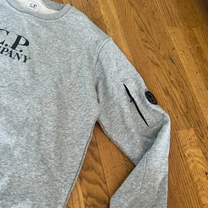 Grå sweatshirt från C.P. Company - Snygg grå sweatshirt från C.P. Company med svart logga på bröstet och ikonisk linsdetalj på ärmen. Tröjan har rund halsringning, lång ärm och en diskret ficka med dragkedja på ärmen. Tillverkad i mjuk bomull för maximal komfort.