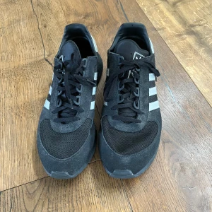 Svarta Adidas sneakers med mesh och mocka - Säljer ett par svarta Adidas sneakers med klassiska vita ränder på sidorna. Skorna har en kombination av mesh och mocka, snörning framtill och en unik sula med rutmönster på sidorna. Perfekta för dig som gillar sportig och stilren look. Storlek 46