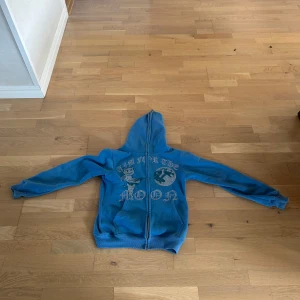 Blå hoodie från Aim For The Moon XS - Blå hoodie med dragkedja från Aim For The Moon i storlek XS. Tryck på framsidan med text och motiv, huva och två fickor framtill. Mjuk insida och långa ärmar, perfekt för chill dagar. Nypris 800kr