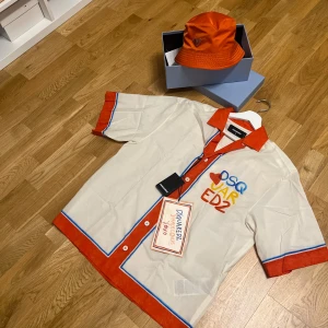 DSQUARED2 kortärmad skjorta orange/vit - Snygg kortärmad skjorta från DSQUARED2 i vit med orange detaljer och blå ränder. Skjortan har färgglad broderad logga på bröstet och knappar framtill. Perfekt för dig som vill sticka ut med en cool och färgstark look.