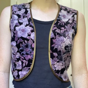 Lila blommig väst med guldkant - Snygg väst med blommigt mönster i lila och svart, kantad med guld. Västen är ärmlös och har en öppen front med rundad nederkant. 