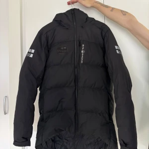 Svart dunjacka från Sail Racing M - Svart dunjacka från Sail Racing med huva och dragkedja framtill. Jackan har loggor på bröstet, ärmarna och huvan. Puffig modell som håller värmen, perfekt för kalla dagar. Snygg och sportig look med diskreta detaljer och hög krage. Kvitto finns!