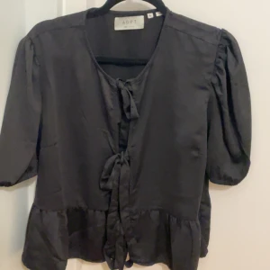 Svart blus med knyt från ADPT - Svart blus från ADPT i storlek XL med kort puffärm och tre dekorativa knyt framtill. Blusen har volangkant nedtill och är tillverkad i ett glansigt, mjukt material som ger en lyxig känsla. Perfekt för dig som gillar trendiga detaljer och en feminin look.