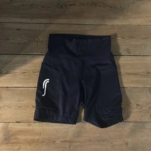 Svarta högmidjade träningsshorts - Snygga svarta högmidjade träningsshorts från Salming med vit logga på benet. De har en tight passform och är gjorda i ett stretchigt syntetmaterial som passar perfekt för gymmet eller löprundan. Praktiska fickor på sidorna för mobilen eller nycklar.