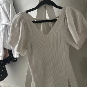 Vit ribbad kortklänning med puffärm - Supersöt vit ribbad klänning med puffiga korta ärmar och v-ringning både fram och bak. Klänningen har knytband i ryggen som ger en extra detalj. Perfekt för dig som gillar en feminin och trendig stil.