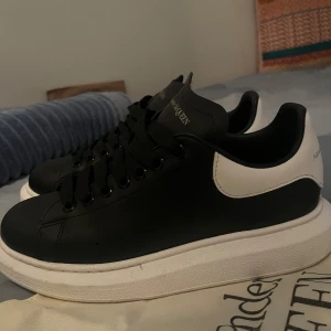 Alexander McQueen svarta sneakers - Svarta sneakers från Alexander McQueen med vit häl och tjock vit sula. Klassisk rund tå, svarta skosnören och logga på plösen och hälen. Tillverkade i skinn för en lyxig känsla och stilren look. Perfekta för dig som gillar statement-sneakers.