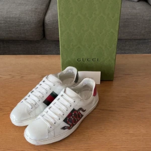 Gucci Ace sneakers med orm och röd detalj - Gucci Ace sneakers i vitt skinn med klassiska gröna och röda ränder på sidan samt en broderad orm i rött och svart. Baksidan har metallicröda och gröna detaljer. Snygg och ikonisk modell med platt sula och snörning, perfekt för dig som vill sticka ut. Storlek 43.5
