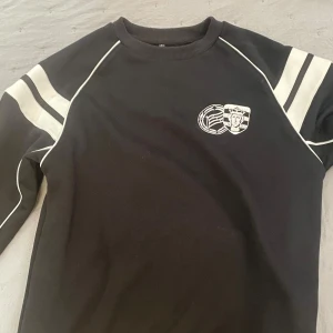 E1 tröja - Svart sweatshirt med vita ränder på ärmarna och vita piping-detaljer längs sömmarna. Tryckt emblem på bröstet med sportig känsla. Tröjan har rund hals och långa ärmar, perfekt för en chill och sportig stil.