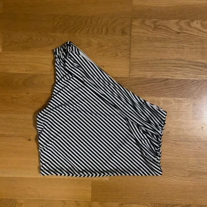 Randig one shoulder topp i svartvitt - Säljer en one shoulder topp med svarta och vita diagonala ränder. Toppen är ärmlös med en smal axelrem på ena sidan och har en figurnära passform. Tillverkad i mjukt och stretchigt material (satin) med vitt innehåll.  Strl 38 men passar 36 också! 