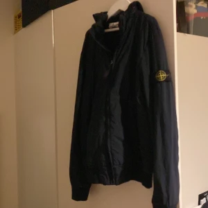 Stone island jacka junior - Storlek 156/12. Köpt för några år sedan. Enda defekt är ett ytterst litet hål under höger arm men syns inte för att det är på undersidan, ifall det är en dealbreaker kan man enkelt fixa ett litet lagningsplåster. Annars utöver hålet är det bra skick. Man kan vika in luvan och ha den utan luva ifall man vill. Äkta såklart. Säljs pga för liten. Perfekt höstjacka för att blockera vinden. STONE ISLAND JUNIOR. Stone island i övrigt är ett lyxigt bra märke. Skriv för frågor om pris eller vara.