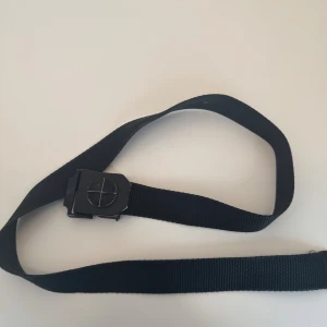 Svart nylonbälte från Stone Island - Säljer ett svart bälte från Stone Island med klassisk kompasslogga på det mattsvarta spännet. Bältet är gjort i slitstark nylon och har en enkel, rak form som passar till jeans eller cargo. Perfekt för dig som gillar streetwear och vill ha en clean look.