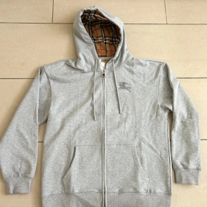 Grå Burberry hoodie med rutig huva - Snygg grå hoodie från Burberry med klassiskt broderat riddarlogo på bröstet och dragkedja framtill. Huvan är fodrad med ikoniskt rutigt Burberry-tyg. Perfekt för dig som vill ha en stilren och lyxig streetwear-look.                 Storlek S men passar även M!                               Pris kan diskuteras!😁