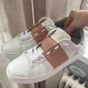 Valentino Garavani Open sneakers vit/rosa - Säljer nu mina snygga Valentino Garavani Open sneakers i vitt skinn med bred ljusrosa läderdetalj över snörningen. Klassisk rund tå och platt sula, samt ikoniska pyramidnitar bak på hälen. Köptes för 6800kr. Använda ca 5 gånger. Box ingår+påse. Skriv om du har frågor!💖