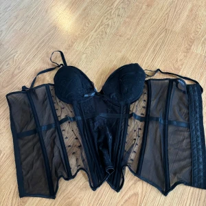 Svart spetskorsett med mesh och hjärtan - Säljer en svart korsett från Toplady med spetsdetaljer och meshpaneler med små hjärtan. Den har vadderade kupor, justerbara axelband och hakknäppning bak. Snyggt markerad midja och transparenta partier för extra edge. Storlek 75B. Använd en gång.