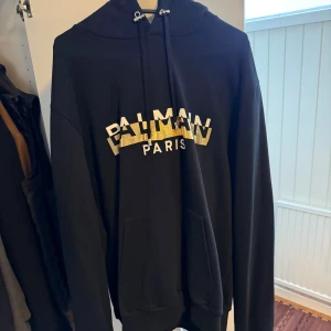 Svart Balmain hoodie med guldtryck - Säljer en svart hoodie från Balmain Paris med stort guldtryck på bröstet. Tröjan har huva med snörning och en stor magficka. Perfekt för dig som vill ha en snygg och trendig streetstyle-look.