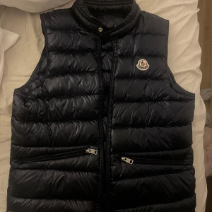 Svart dunväst från Moncler - Snygg svart dunväst från Moncler med klassisk quiltad design och hög krage. Västen har två dragkedjefickor framtill och Monclers ikoniska logga på bröstet. Perfekt för lager-på-lager och streetstyle. Dragkedjan är lite svår.