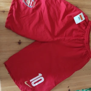 Arsenal röda fotbollsshorts Nike - Röda Arsenal-shorts från Nike med broderat klubbmärke, tryckt nummer 10 och stor vit Arsenal-text på baksidan. Klassisk sportig look med resår i midjan, perfekt för fotbollsplanen. Materialet är lätt och andas, vilket gör dem sköna att spela i.