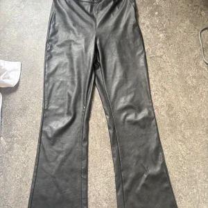 Svarta bootcut byxor i skinnlook Gina Tricot - Snygga svarta byxor i skinnimitation från Gina Tricot. Modellen har bootcut-ben och dragkedja framtill. Perfekta för dig som vill ha en edgy vibe och gillar trendiga detaljer. Passar till allt från sneakers till boots.