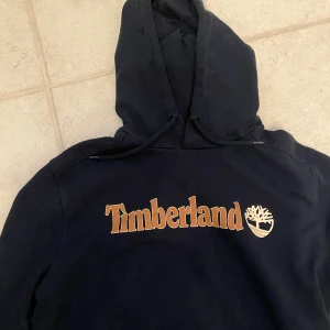 Mörkblå Timberland hoodie med logga - Säljer en mörkblå hoodie från Timberland med stor logga i beige framtill. Tröjan har huva med snörning och är tillverkad i en mjuk bomulls- och polyestermix. Regular fit och perfekt för chill dagar.