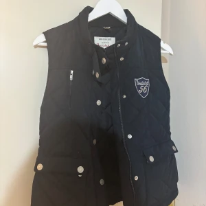 Svart quiltad dunväst från Bondelid - Svart quiltad dunväst från Bondelid med hög krage, silverfärgade knappar och dragkedja. Västen har två fickor med lock och en bröstficka med dragkedja samt ett broderat märke på bröstet. Perfekt för lager-på-lager under kyliga dagar.