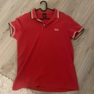 Röd pikétröja från BOSS Hugo Boss - Röd pikétröja från BOSS Hugo Boss med vit logga på bröstet. Klassisk krage med vita och svarta ränder samt matchande detaljer på ärmsluten. Skön passform och stilren look, perfekt för dig som gillar snygga basplagg.