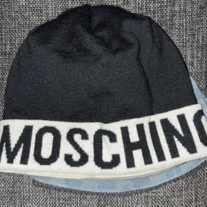 Svart och vit Moschino mössa - Snygg svart mössa från Moschino med bred vit kant där Moschino-loggan är stickad i svart. Mössan är tillverkad i mjuk polyester och har en klassisk, rundad form som sitter skönt på huvudet. Perfekt för dig som gillar streetstyle och vill sticka ut med ett designerplagg.