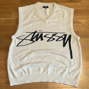 Gräddvit stickad väst från Stüssy - Säljer en vit stickad väst från Stüssy i 100% bomull. Västen har en v-ringad hals och ett stort svart Stüssy-logotryck över bröstet. Ribbad kant vid ärmhål, hals och nederkant. Perfekt för lager-på-lager och streetwear-stil.