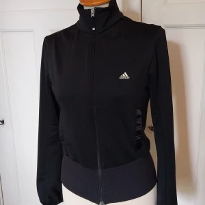 Svart träningsjacka från Adidas M - Svart träningsjacka från Adidas med hel dragkedja, högt krage och stora loggor både fram och bak. Jackan är i mjukt och stretchigt material, har ribbade muddar och Clima365-teknologi för extra komfort. Perfekt för dig som gillar sportig stil.