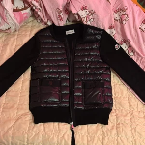 Svart moncler cardigan  - Svart pufferjacka från Moncler med quiltad framsida och två fickor med knappstängning. Jackan har långärmade släta ärmar och ribbad mudd vid midja och ärmslut. Klassisk Moncler-logga på ena ärmen och dragkedja framtill. Perfekt för kyliga dagar. (Använts 3 gånger)