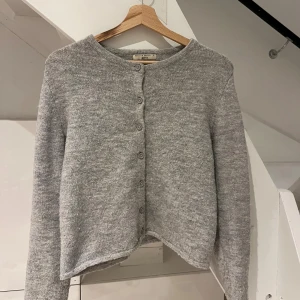 Grå kofta från Gina Tricot - Mysig ljusgrå kofta från Gina Tricot i stickad ullmix. Klassisk rak modell med rund halsringning och knappar framtill. Perfekt att slänga över ett linne eller t-shirt för extra värme. Långärmad och enkel att matcha till allt.