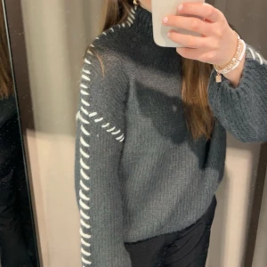 Grå stickad tröja från Vila - Mysig grå stickad tröja från Vila med vita dekorativa detaljer längs ärmar och axlar. Tröjan har en hög hals och lång ärm, perfekt för kyliga dagar. Den har en loose fit och känns både trendig och bekväm.