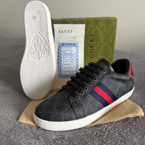 Gucci skor  - Snygga Gucci sneakers i grå/svart med GG-monogram över hela skon. Sidorna har klassiska röd-blå ränder och hälarna är i matallic röd och blå reptilstruktur med Gucci-logga. Skorna har snörning, platt vit sula och rund tå. Materialet är canvas och skinn.