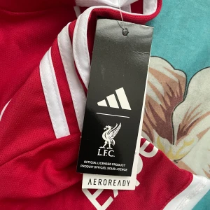 Liverpool FC röd matchtröja Adidas - Liverpool FC officiell matchtröja från Adidas med tryck 'ISAK 9' på ryggen. Röd med vita detaljer, korta ärmar och klassiska Adidas-ränder på axlarna. Materialet är lätt och ventilerande Aeroready-polyester. UEFA Foundation-märke på ärmen och Standard Chartered-logga på bröstet.