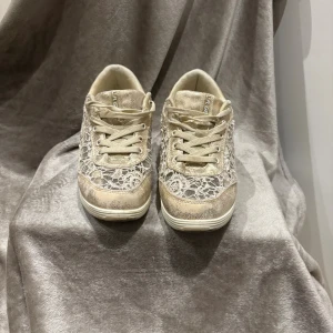 Guldiga sneakers med spets från LA Gear - Unika sneakers från LA Gear i guldigt syntetmaterial med vita spetsdetaljer och snörning. Skorna har en chunky vit sula och en metallic guldhäll på hälen som ger extra bling. Perfekta för dig som vill sticka ut med en cool och feminin stil.