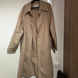 Beige trenchcoat från Levate - Stilren beige trenchcoat från Levate med klassisk krage och knäppning framtill. Jackan har långa ärmar och en lös passform.