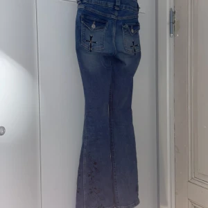 Blå bootcut jeans med broderi - Unika blå jeans med bootcut passform och snygga broderade blommor längs benen. Bakfickorna har coola korsdetaljer och knappar. Längst ner på benen är broderade blommor med en själkdesign.