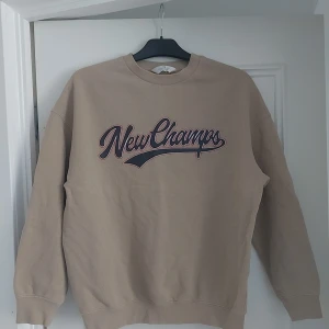 Beige sweatshirt från H&M med tryck 12-14 år - Beige sweatshirt från H&M med coolt 'New Champs'-tryck i mörkblått och vinrött på bröstet. Tröjan har rund hals, ribbade muddar och är långärmad. Perfekt för en avslappnad och trendig stil.