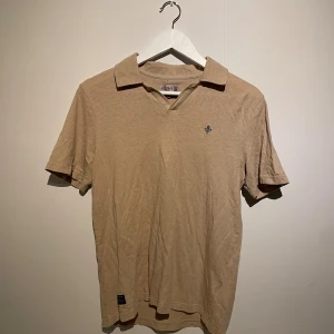 Beige t-shirt piké med krage från Morris - Säljer en beige t-shirt från Morris i storlek S. T-shirten har klassisk krage, korta ärmar och en liten broderad logga på bröstet. Materialet är mjuk bomull och färgen är neutral beige, perfekt för en clean och chill look.